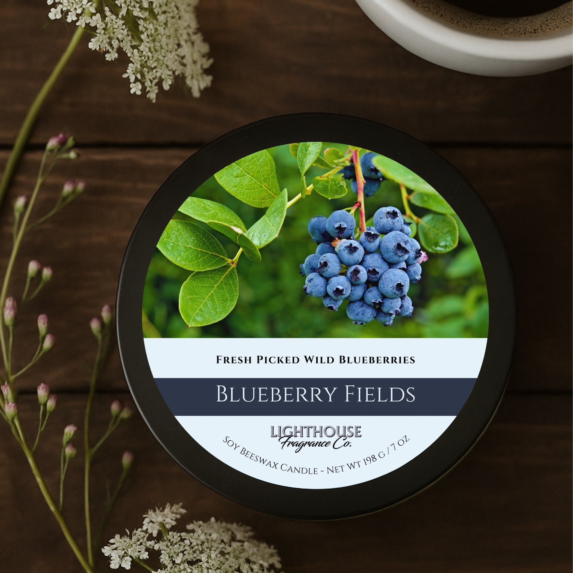 Candle labeled 'Blueberry Fields' by Lighthouse Fragrance Co. on a wooden surface with flowers.