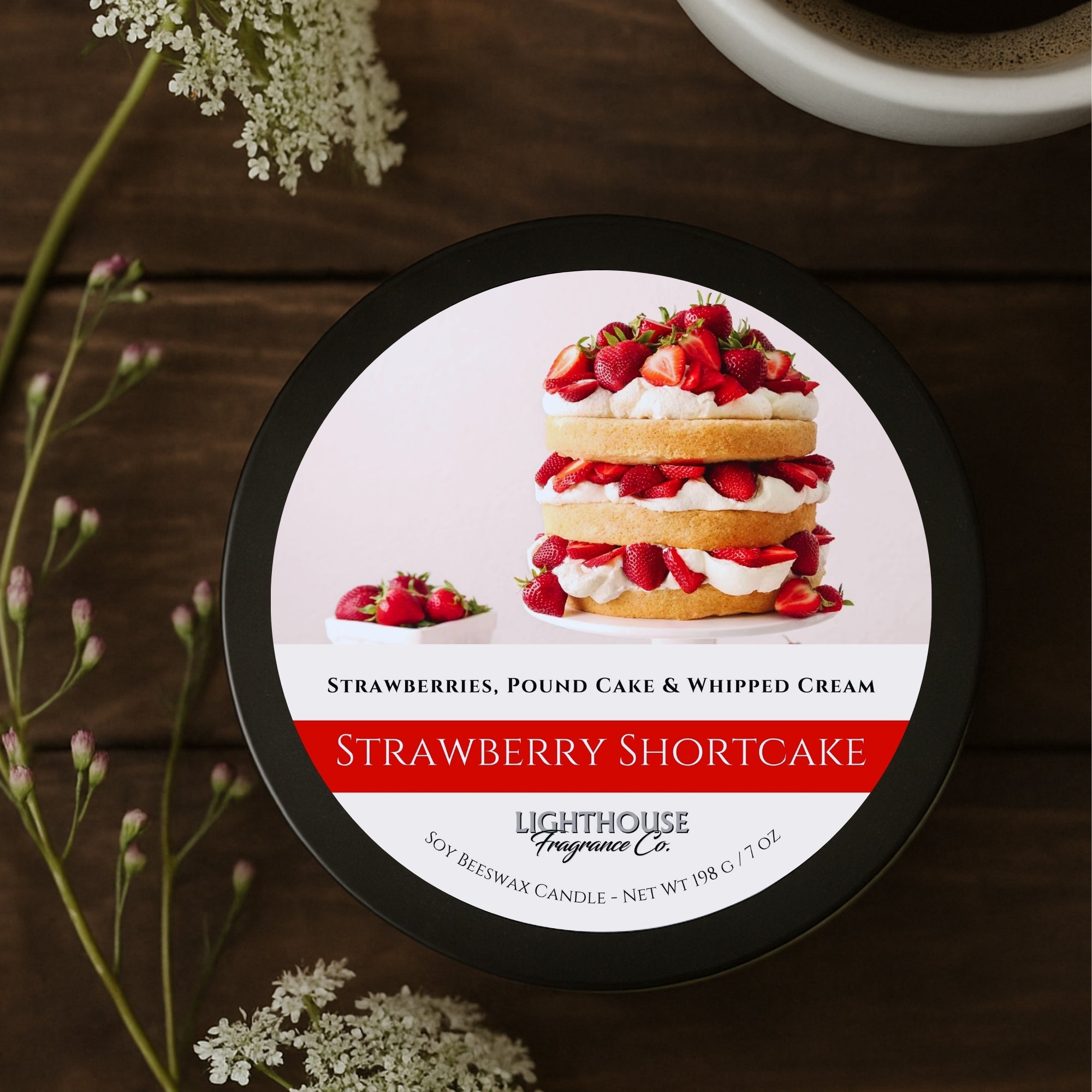 Candle labeled 'Strawberry Shortcake' from Lighthouse Fragrance Co. on a wooden surface with flowers.
