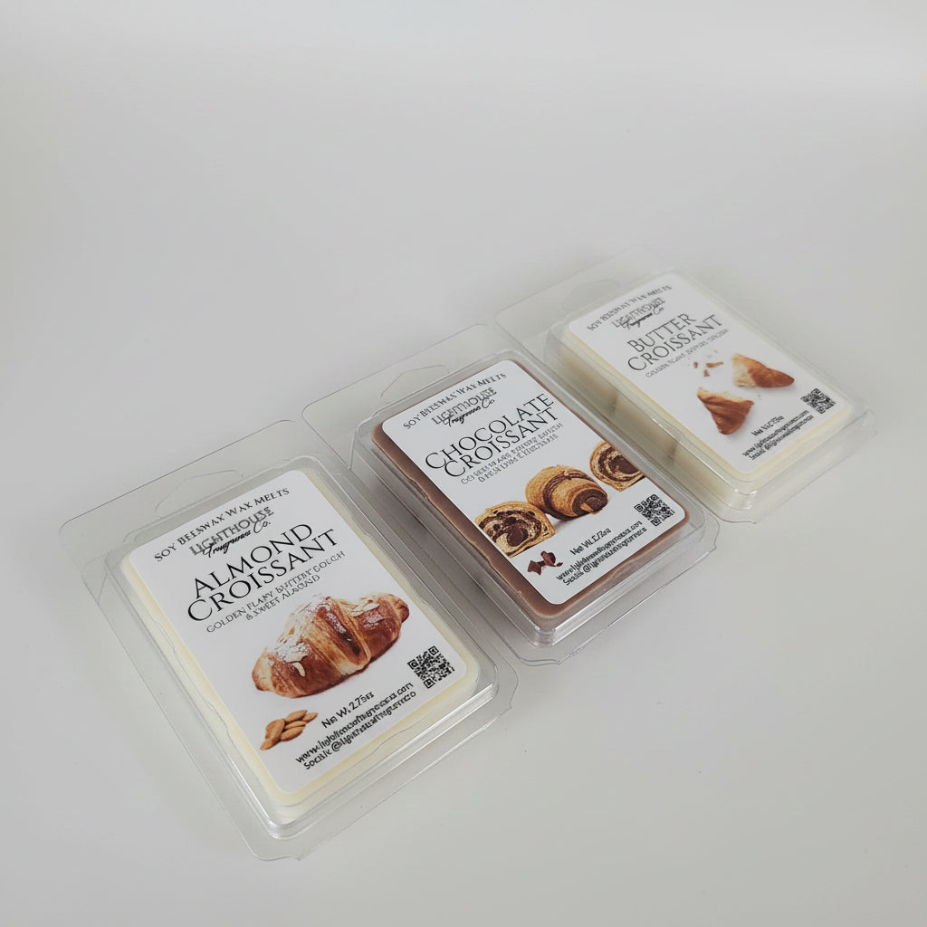 Three packaged wax melts in clear plastic clamshell containers arranged on a stainless steel surface. The front two packages are labeled Almond Croissant and Chocolate Croissant, and the back two packages are Butter Croissant.