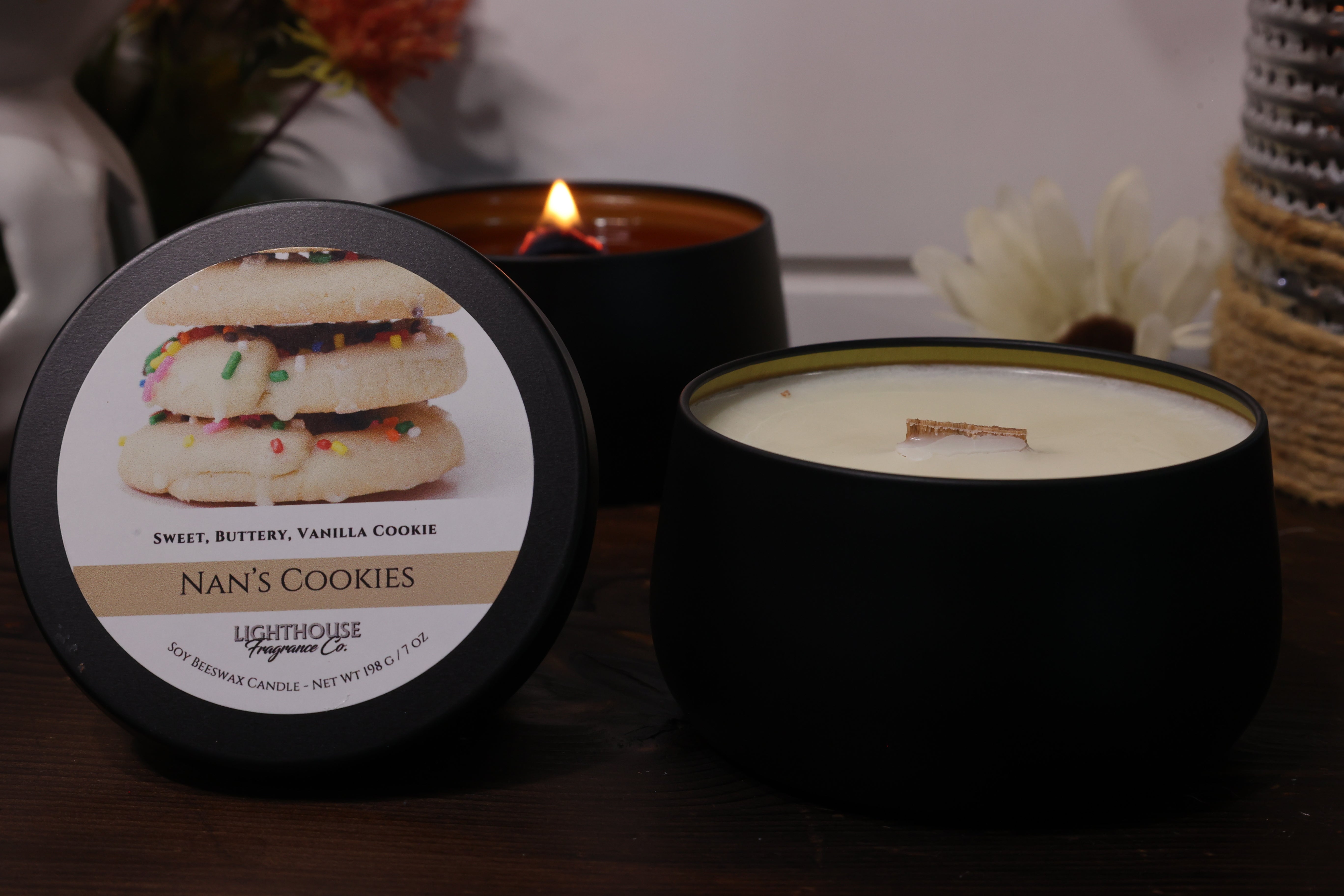 Two candles, one lit and one unlit, with a container labeled 'Nan's Cookies' on a wooden surface.