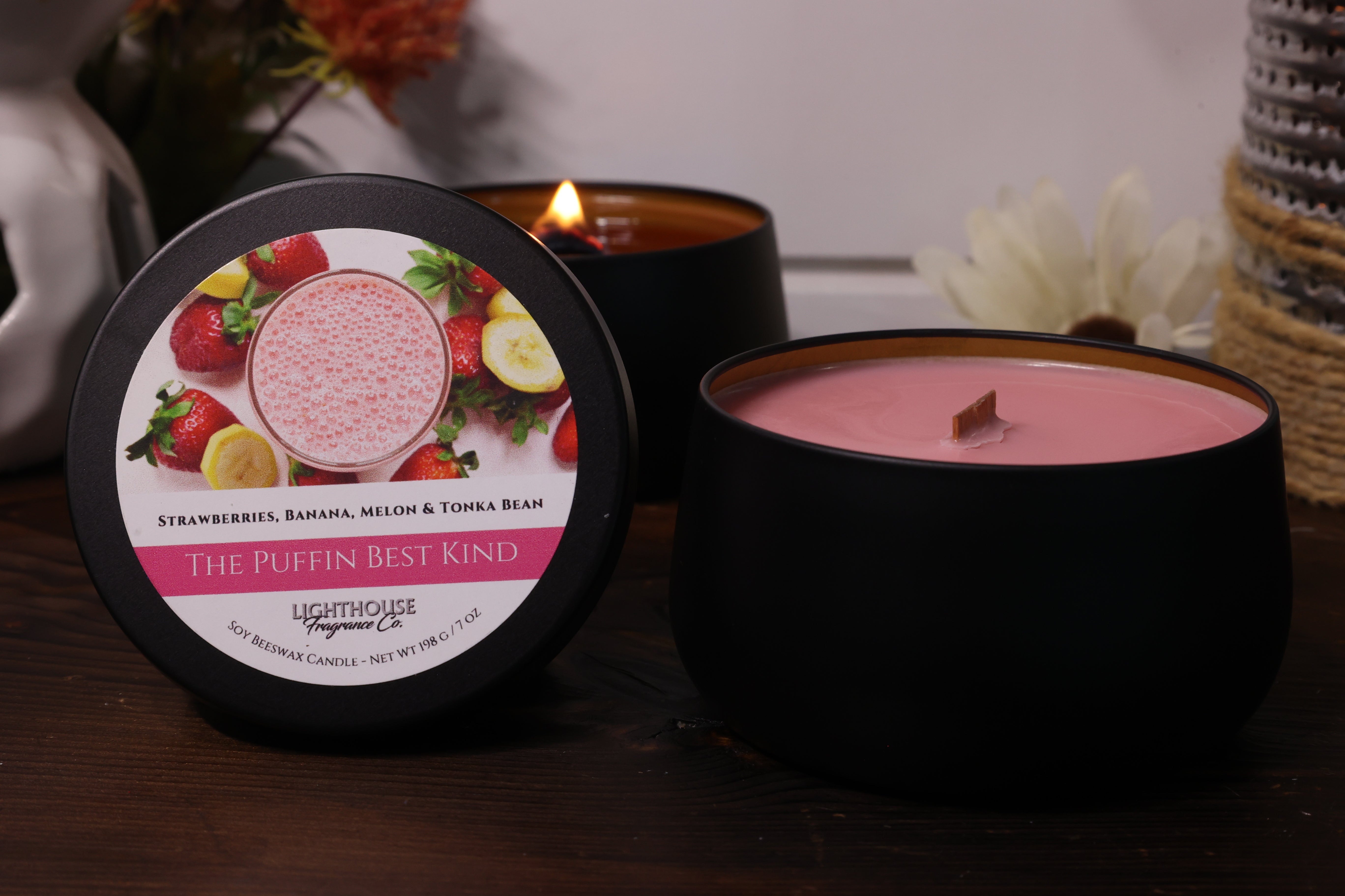 Pink candle in a black container with a decorative label on a wooden surface.